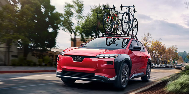 Red 2026 Subaru Solterra electric SUV driving through a city street with bicycles mounted on the roof rack.