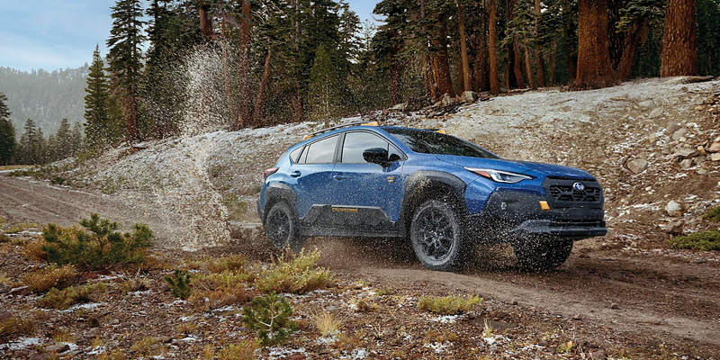 Blue 2026 Subaru Crosstrek driving off road on a muddy forest trail with water splashing around the tires.