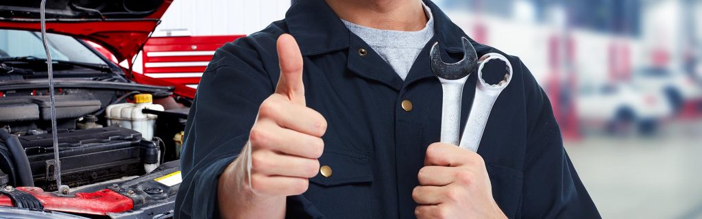 A mechanic giving a thumbs up while holding two wrenches, with a car engine in the background.